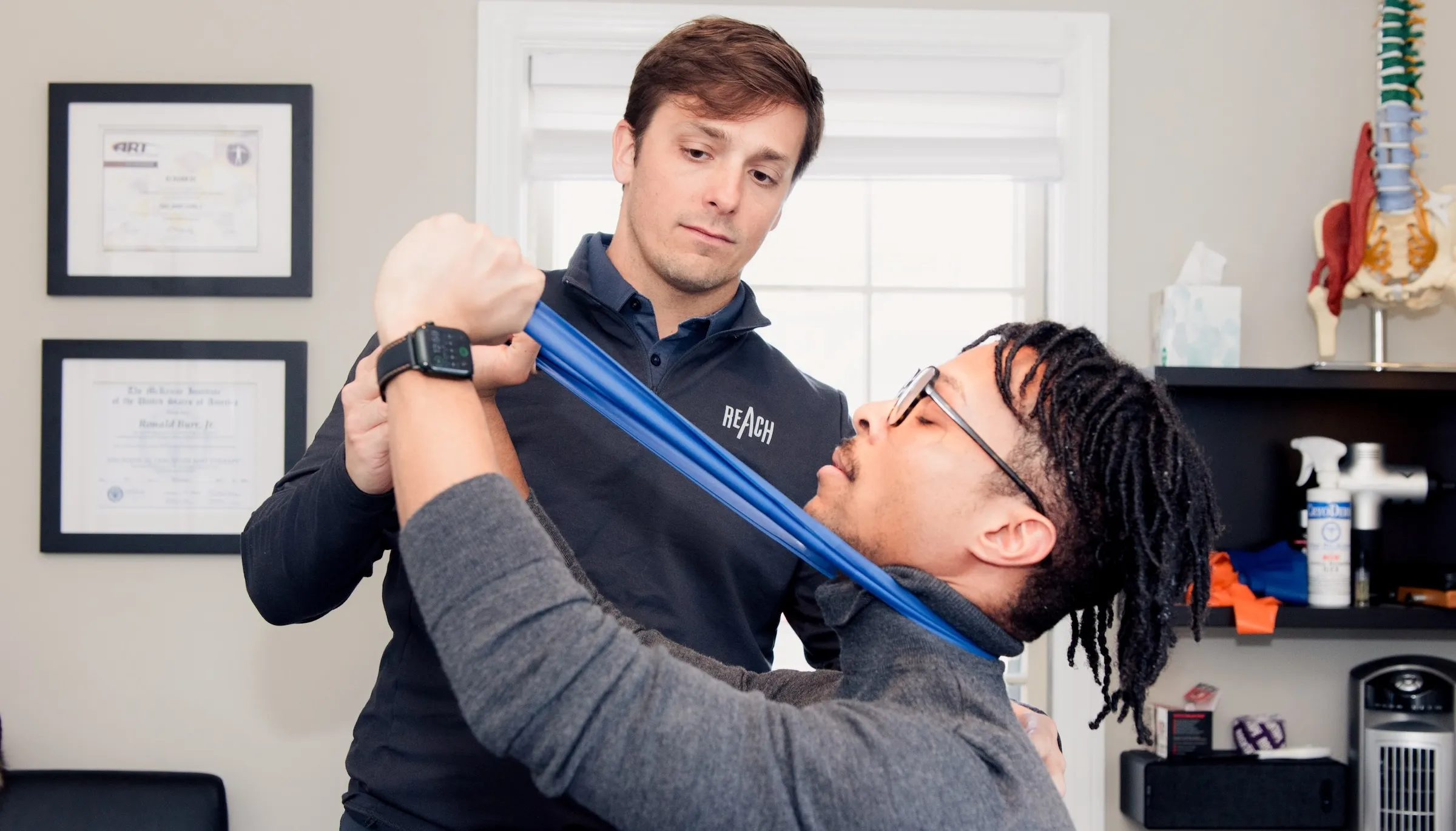Dr. RJ Burr supervising a seated patient performing a banded McKenzie neck retraction exercise at REACH Rehab + Chiropractic, Plymouth MI