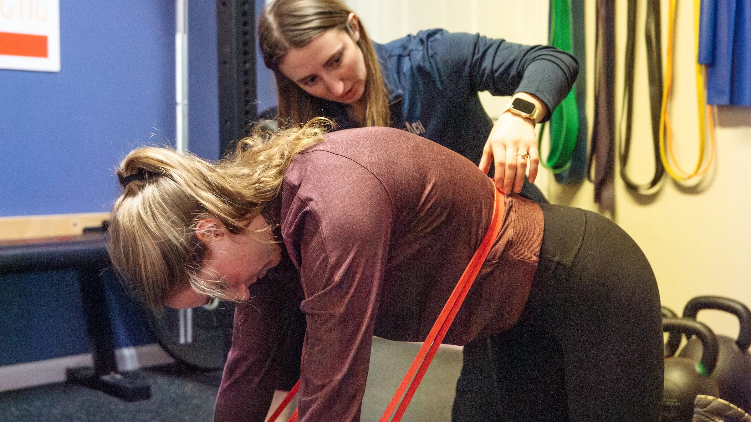 Dr. Sheridan Kent cueing a patient's hip hinge with a resistance band around the hips and a hand on the low back, at REACH Rehab + Chiropractic in Plymouth, MI