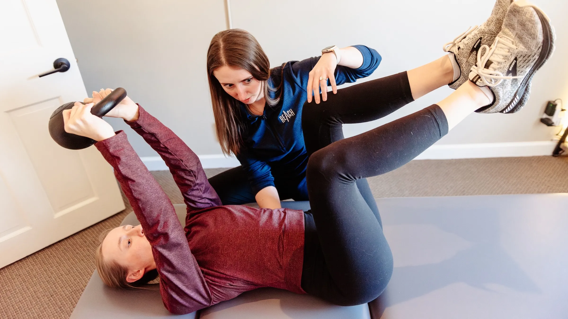 Dr. Sheridan Kent guiding a patient through a DNS dead-bug, holding a kettlebell overhead with hips and knees bent to ninety degrees, at REACH Rehab + Chiropractic in Plymouth, MI