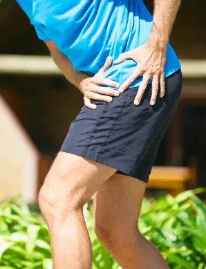 A man outdoors in a blue shirt and dark shorts gripping the outside of his hip, where IT band pain is typically felt