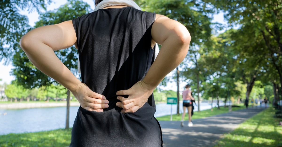 A Runner in a Black Shirt and White Towel Around Their Neck Grabbing This Lower Back in Pain | Going Beyond Back Pain Relief | REACH Rehab + Chiropractic