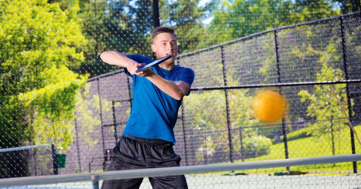 A Man Hitting a Pickleball Over The Net on a Pickleball Court with Fences and Trees in the Background | Pickle Elbow No More: Proven Strategies for Healing and Prevention | REACH Rehab + Chiropractic