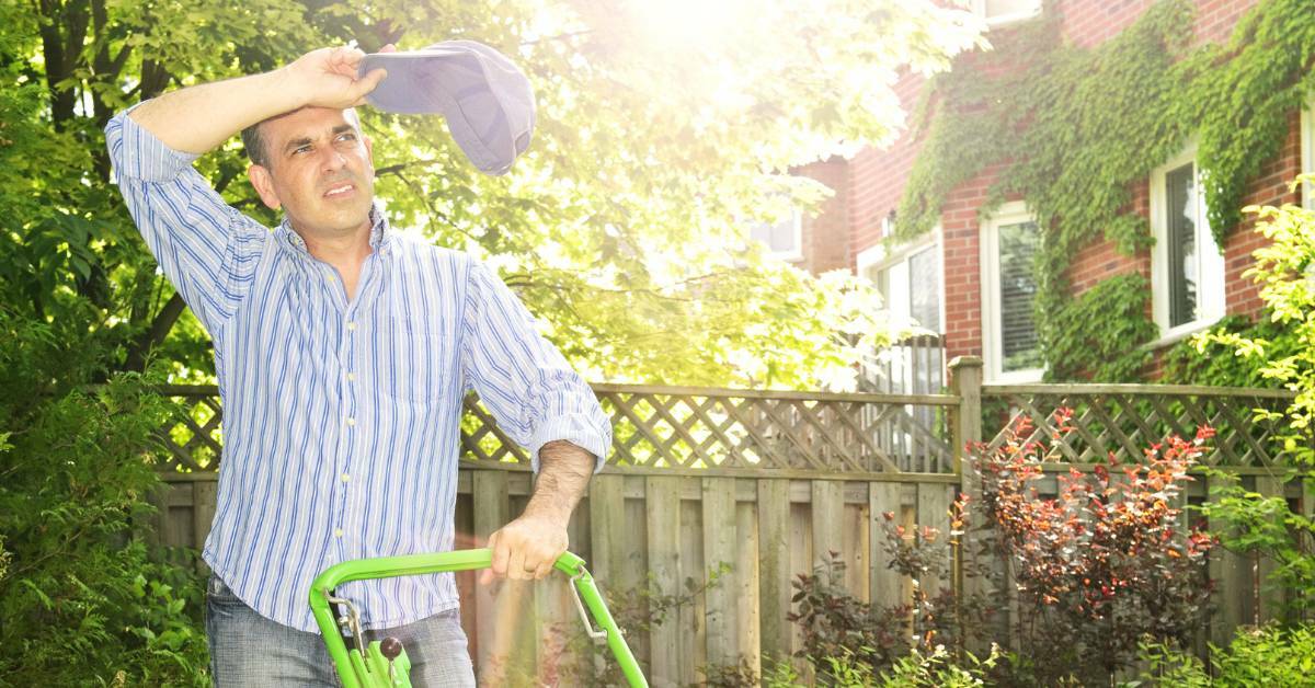 A Man Wiping His Forehead With A Baseball Cap In His Hand While Mowing The Lawn With A Fence and House in The Background | Prevent Injuries During Warm Weather Chores | REACH Rehab + Chiropractic