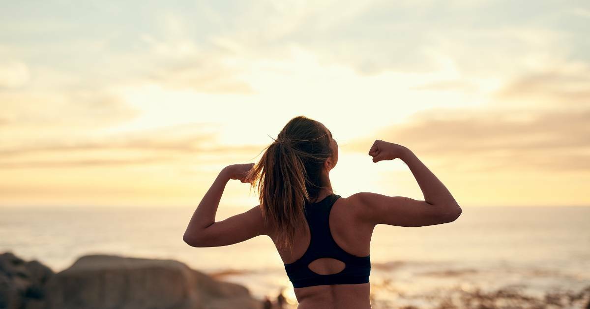 A Woman With Her Back to Us Putting Her Arms in the Air Flexing Her Muscles With a Body of Water and Sunset in the Background | Healthy Summer Habits | REACH Rehab + Chiropractic