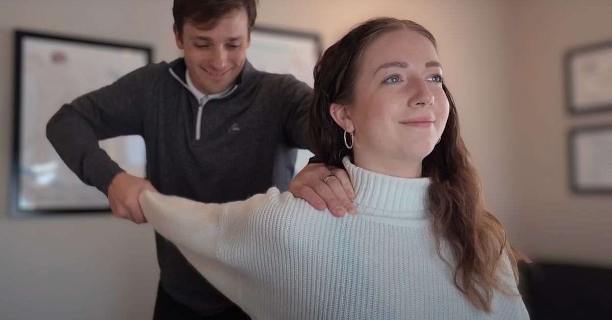 A man assists a smiling woman with an arm and shoulder movement in a professional setting, conveying care and positive interaction.