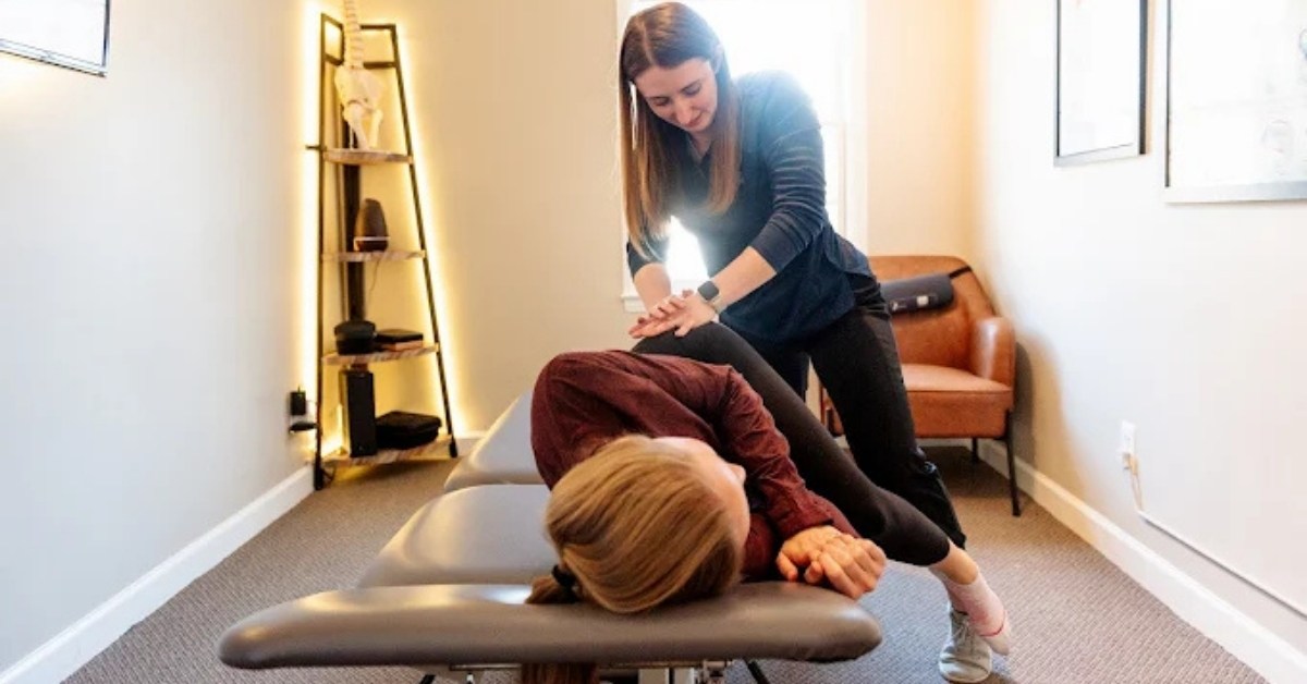 A female chiropractor performs a hip or lower back Cox flexion-distraction adjustment on a patient lying on a grey treatment table in a modern therapy room. The room features a light-colored wall, a dark-framed shelving unit with an anatomical skeleton model, and a tan armchair. The setting is clean, well-lit, and professional.