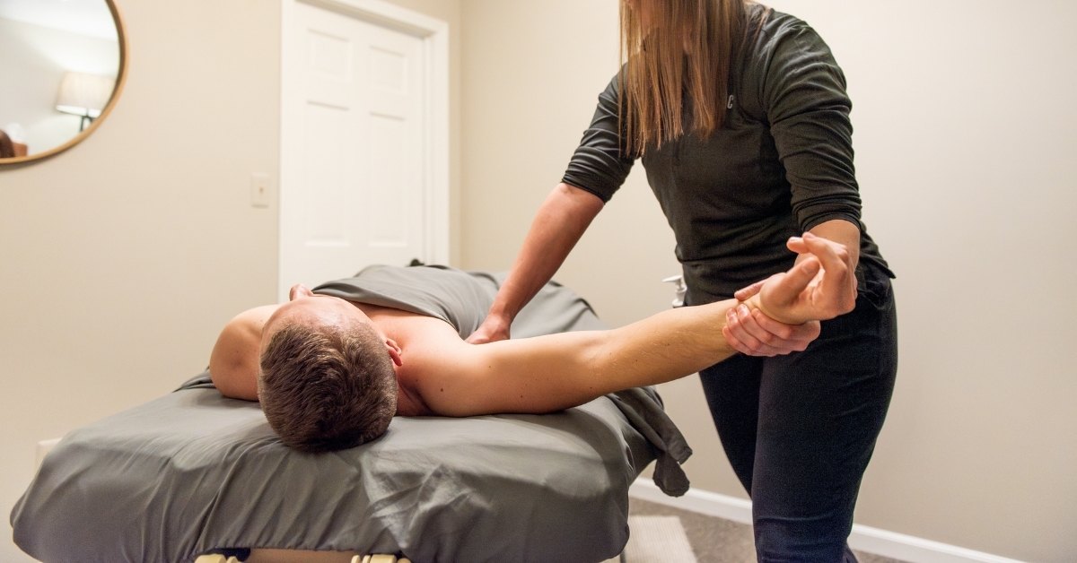 A male client lies face down on a massage table, covered with a gray sheet, while a female therapist performs a manual therapy technique on his right shoulder and arm. The setting is a clean, well-lit therapy room with neutral-colored walls and a round wooden-framed mirror in the background.