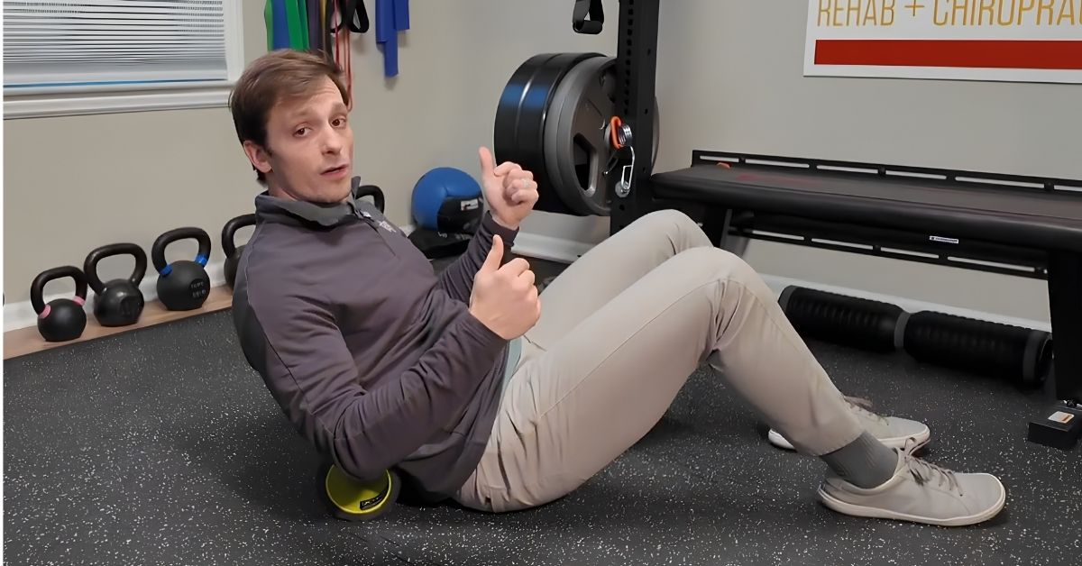 Dr. RJ Burr, REACH Rehab + Chiropractic, sitting on a gym floor using a foam roller under his lower back and giving two thumbs up, surrounded by kettlebells, resistance bands, weights, and a bench in a rehab or chiropractic clinic.